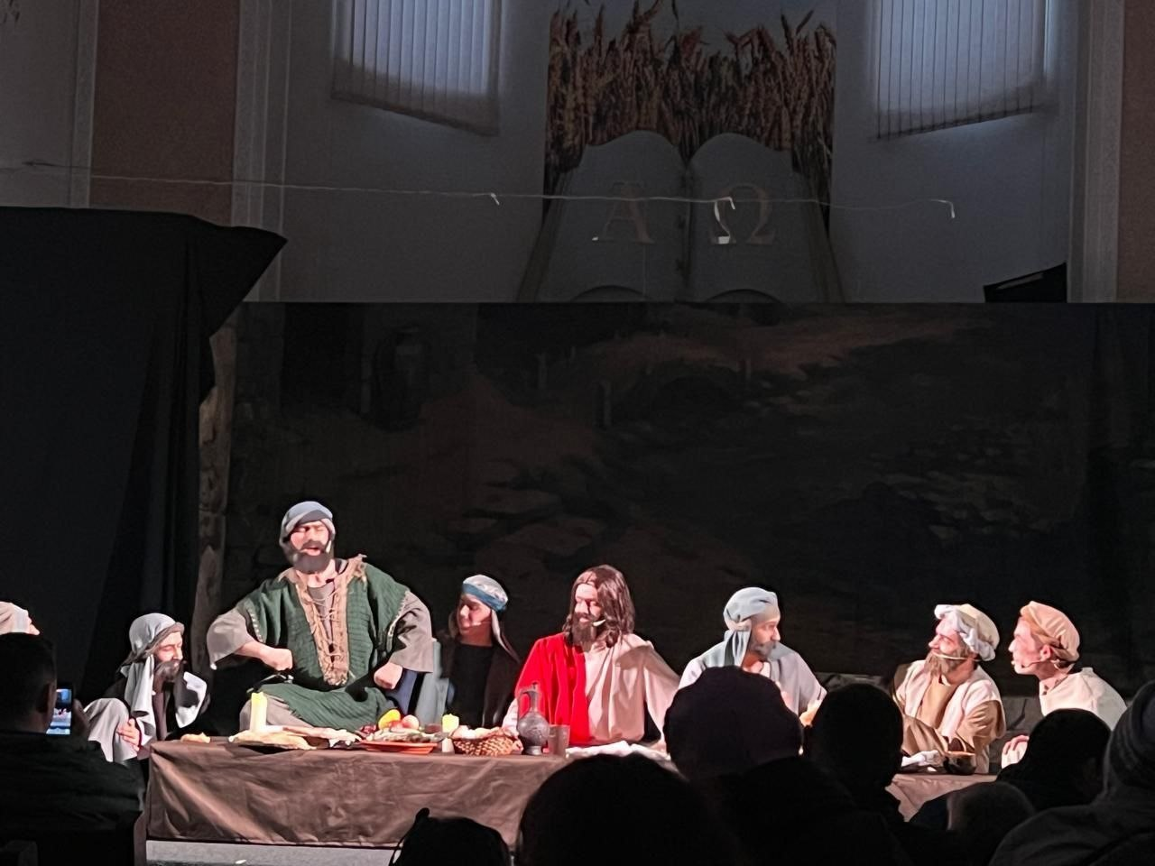 Театральна сцена, що відтворює біблійний сюжет «Таємна вечеря». На сцені за довгим столом сидять актори в історичному вбранні та головних уборах, а глядачі спостерігають за виставою із зали.