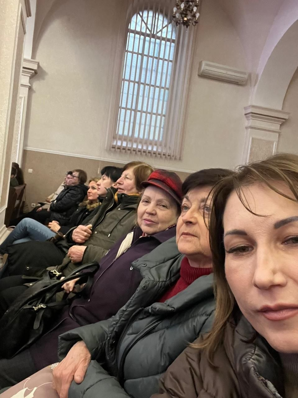 Група жінок різного віку сидить у залі з високими стелями та великим вікном із вертикальними жалюзі. Вони одягнені у верхній одяг і чекають на захід або дивляться виставу, а жінка на передньому плані робить спільне селфі.