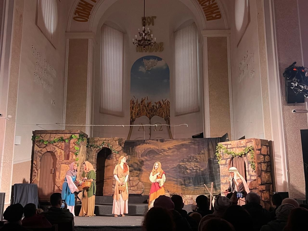 Вистава у великій залі з високим склепінням та релігійною символікою на стіні. На сцені, оформленій декораціями у вигляді кам'яних будівель, актори в автентичному біблійному вбранні розігрують сцену перед глядачами, які сидять у залі.