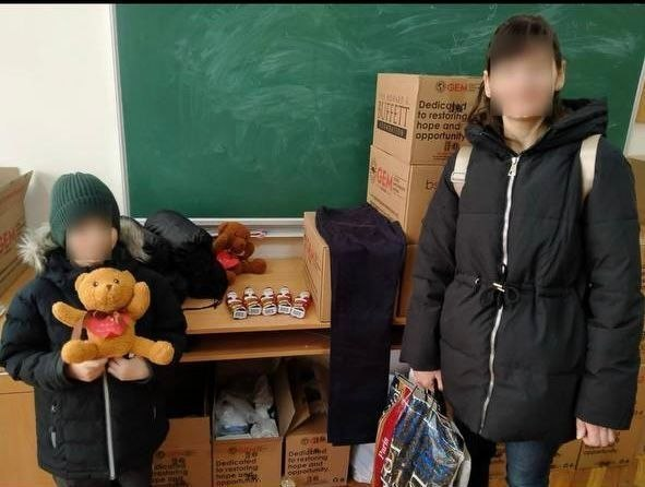 Жінка та дитина в зимовому одязі стоять у навчальному класі поруч із коробками гуманітарної допомоги. Дитина тримає плюшевого ведмедика, а обличчя обох осіб приховані (розмиті) з метою безпеки та анонімності.
