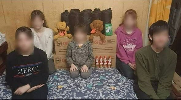 П'ятеро дітей та підлітків сидять на ліжку на фоні коробок із гуманітарною допомогою, рюкзаків та м'яких іграшок. Обличчя всіх присутніх приховані (розмиті) для захисту їхньої приватності.