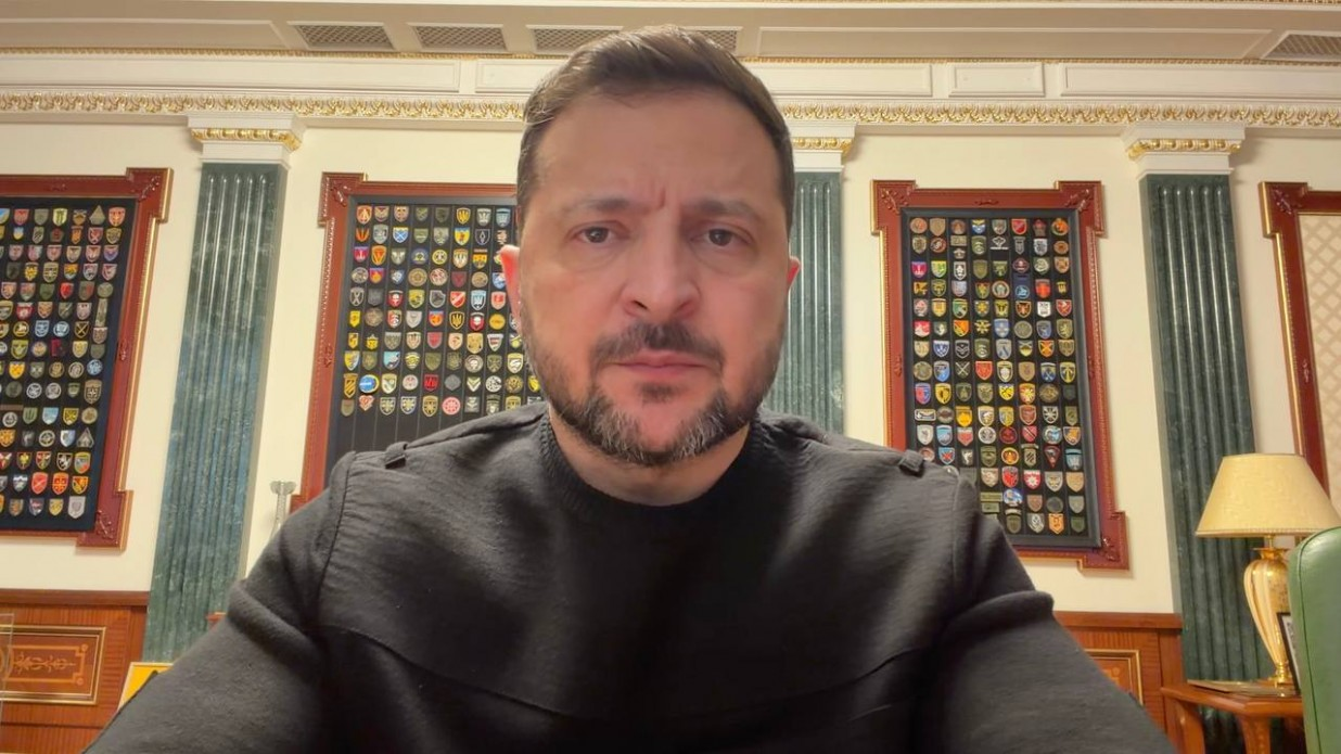 Чоловік у чорному світшоті дивиться прямо в камеру на фоні офісного інтер'єру. За його спиною на стіні розміщені стенди з великою кількістю військових шевронів, а збоку видно настільну лампу та частину класичних меблів.