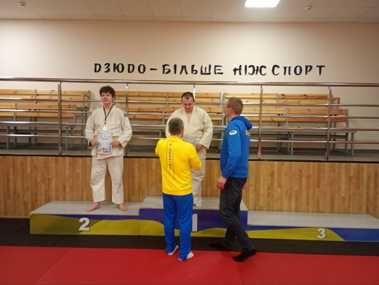 Нагородження призерів змагань із дзюдо. Двоє спортсменів у кімоно стоять на п'єдесталі з медалями та грамотами. Поруч — тренери у спортивній формі. На стіні напис: «Дзюдо — більше ніж спорт».