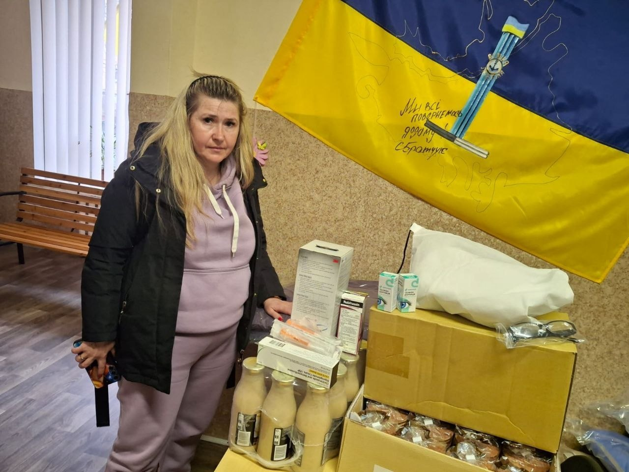 Жінка стоїть поруч із гуманітарною допомогою: коробками з медикаментами, засобами гігієни та продуктами харчування. На стіні позаду розміщено український прапор із підписами та військовою символікою.