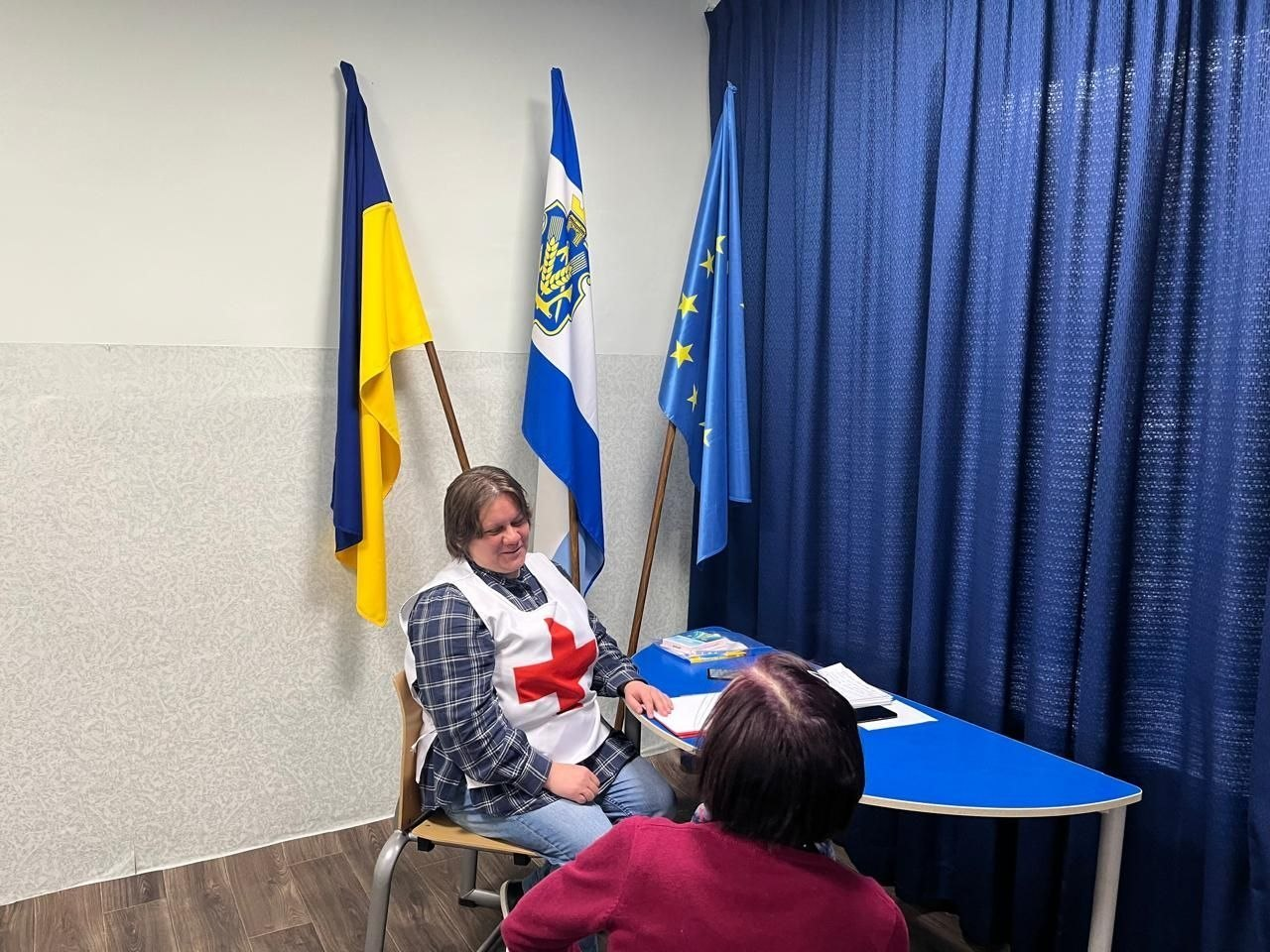 Представниця Товариства Червоного Хреста проводить консультацію для жінки в офісному приміщенні. Позаду них встановлені прапори України, Херсонської області та Європейського Союзу.