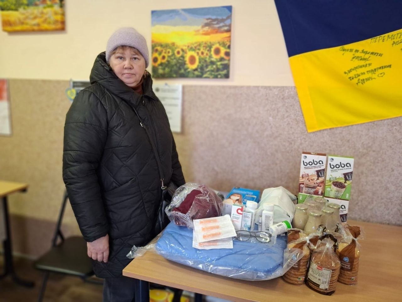 Жінка стоїть біля столу з отриманою гуманітарною допомогою: продуктами харчування, ліками та теплими речами. На стіні позаду розміщені картини з соняшниками та прапор України з побажаннями.