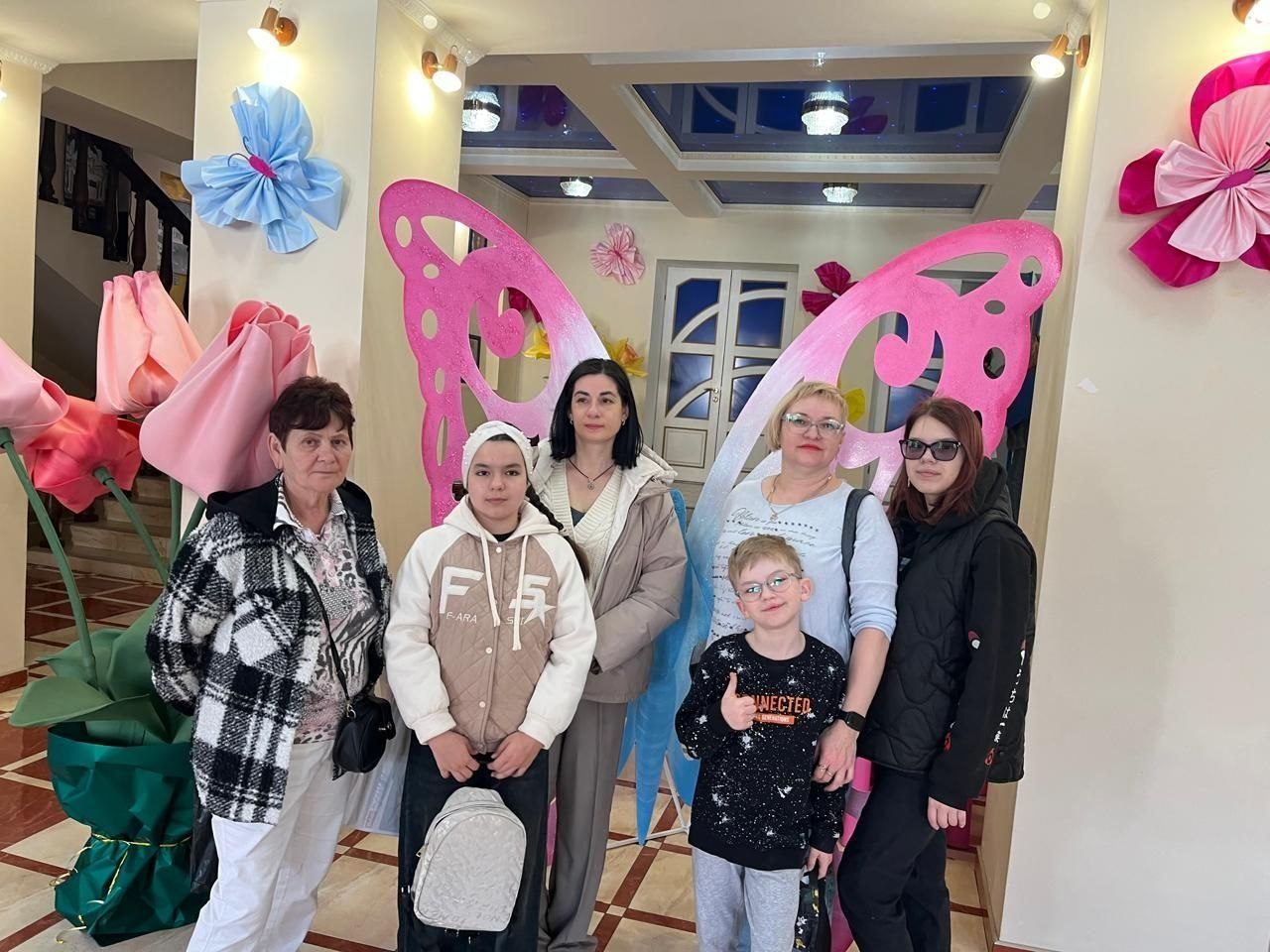 Група людей — четверо жінок та двоє дітей — позують для фото у святково оформленому приміщенні. На фоні розташовані великі декоративні метелики рожевого та блакитного кольорів, а також об’ємні паперові квіти на стінах і колонах.