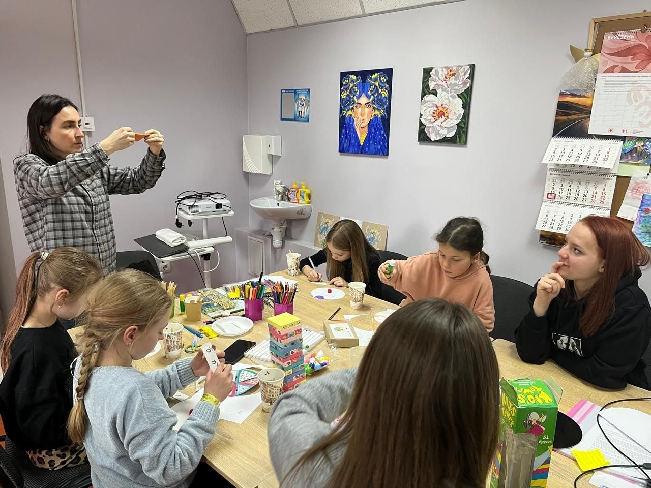 Діти та дорослі займаються творчістю за великим столом у світлій кімнаті. Учасники майстер-класу малюють та розглядають свої роботи, на столі лежать канцтовари, стаканчики та настільні ігри. На стіні позаду висять картини та настінний календар.