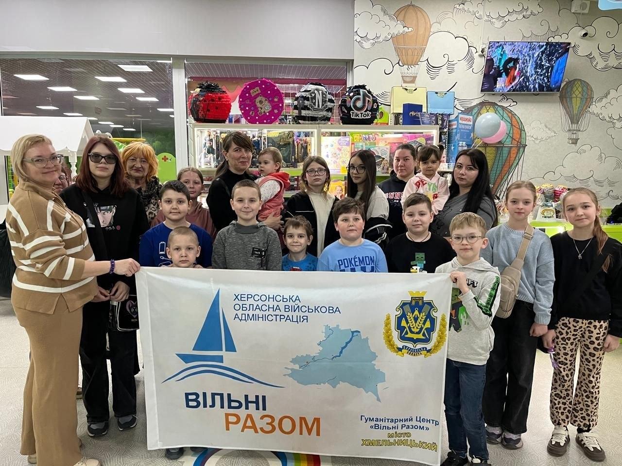 Група дітей та дорослих тримають білий банер із логотипом Херсонської ОВА та написом «Вільні Разом». Вони позують у дитячому ігровому центрі з іграшками на полицях та малюнками повітряних куль на стінах.