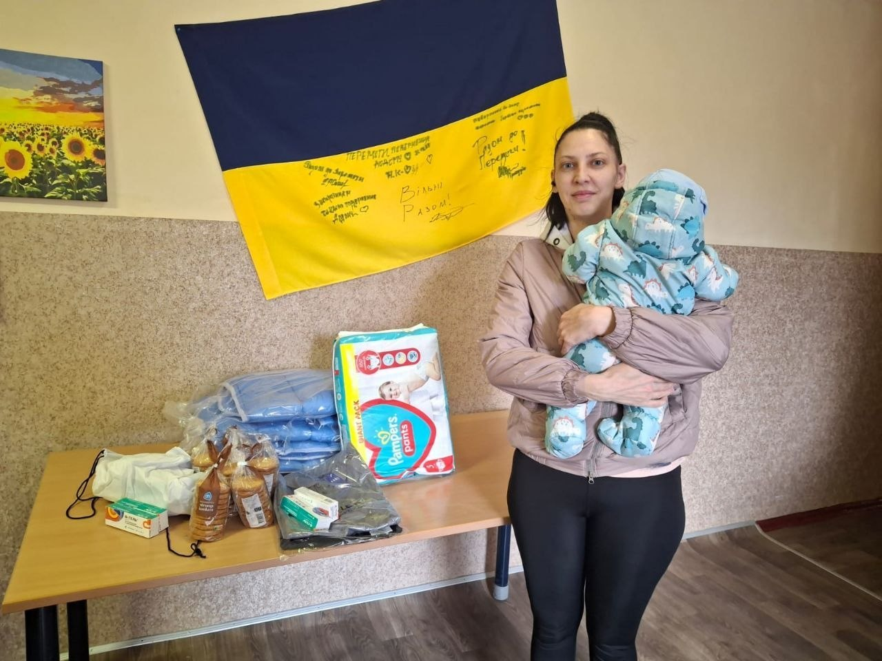 Жінка з немовлям на руках стоїть біля столу з отриманою гуманітарною допомогою: підгузками, ковдрами, медикаментами та продуктами харчування. На стіні позаду розміщено синьо-жовтий прапор із підписами та побажаннями, а поруч — картина із соняшниками.