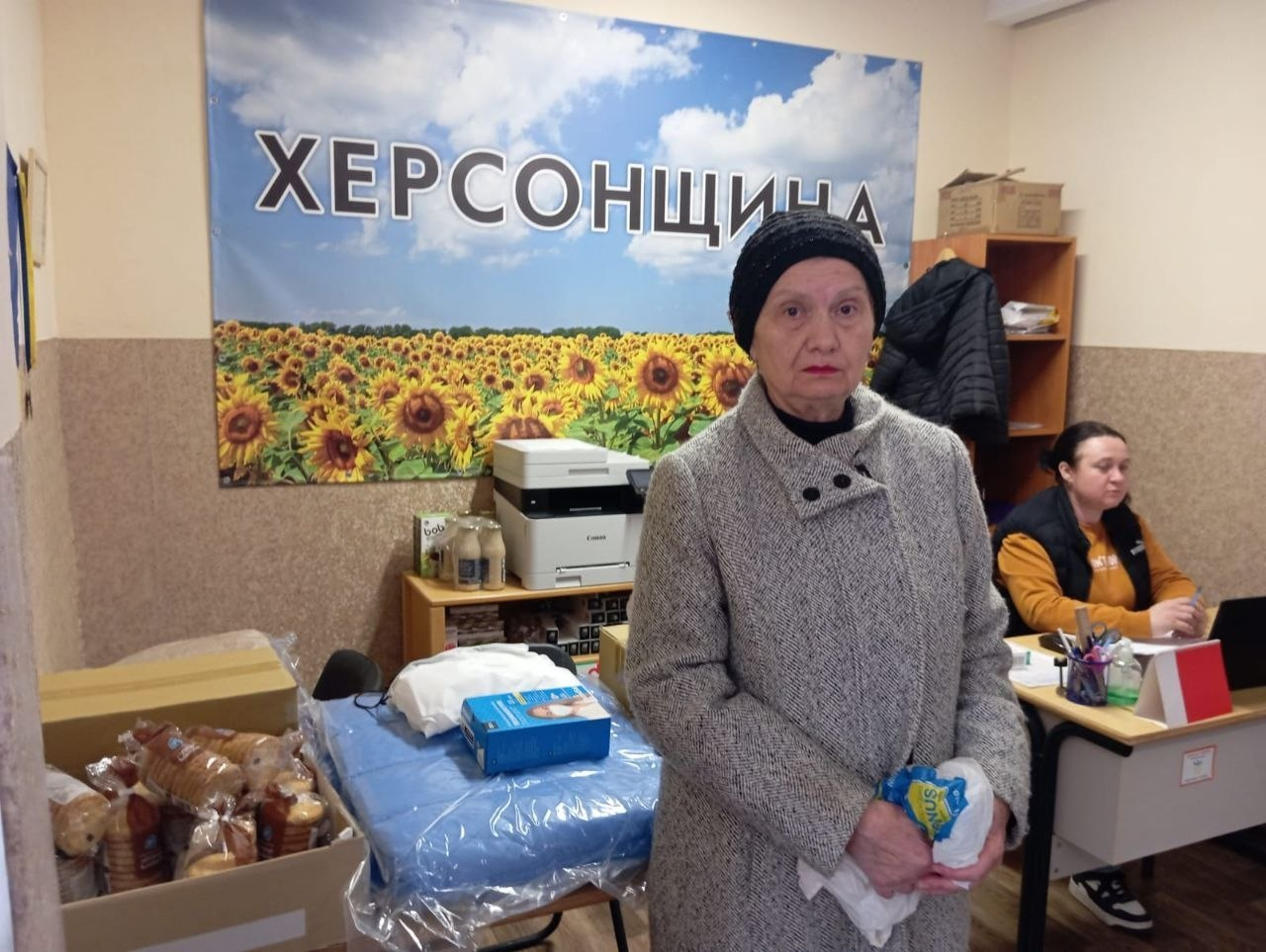 Літня жінка стоїть у приміщенні поруч із гуманітарною допомогою: продуктами харчування, медичними засобами та засобами гігієни. На стіні позаду розміщено великий банер із соняшниковим полем і написом «ХЕРСОНЩИНА».