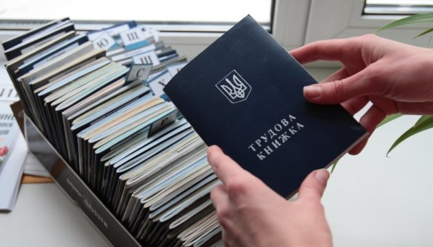 Руки тримають синю трудову книжку з гербом України над органайзером, у якому щільно розставлено багато аналогічних документів із літерними розділювачами.