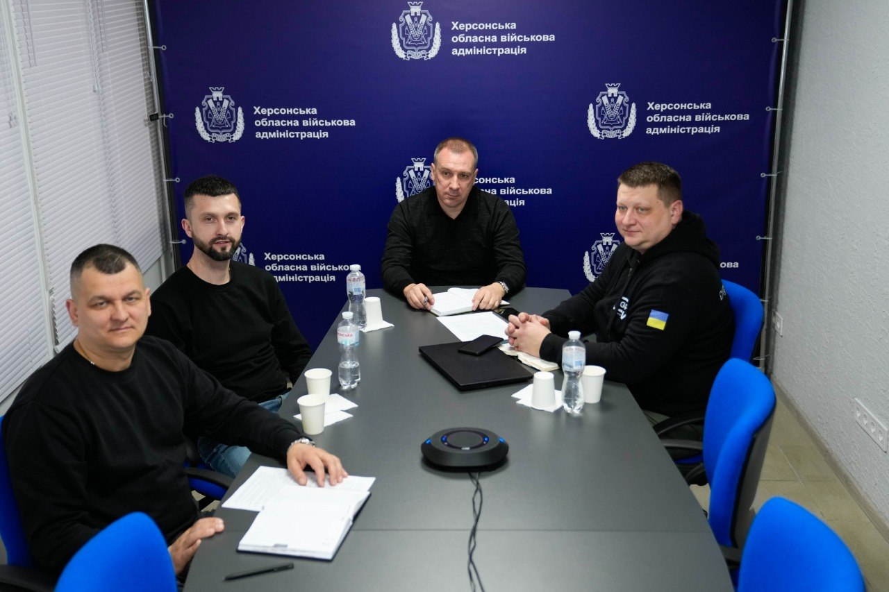 Четверо чоловіків за столом під час робочої наради. Перед ними лежать документи, блокноти та пляшки з водою. Позаду — темно-синій банер із офіційною символікою та гербом.