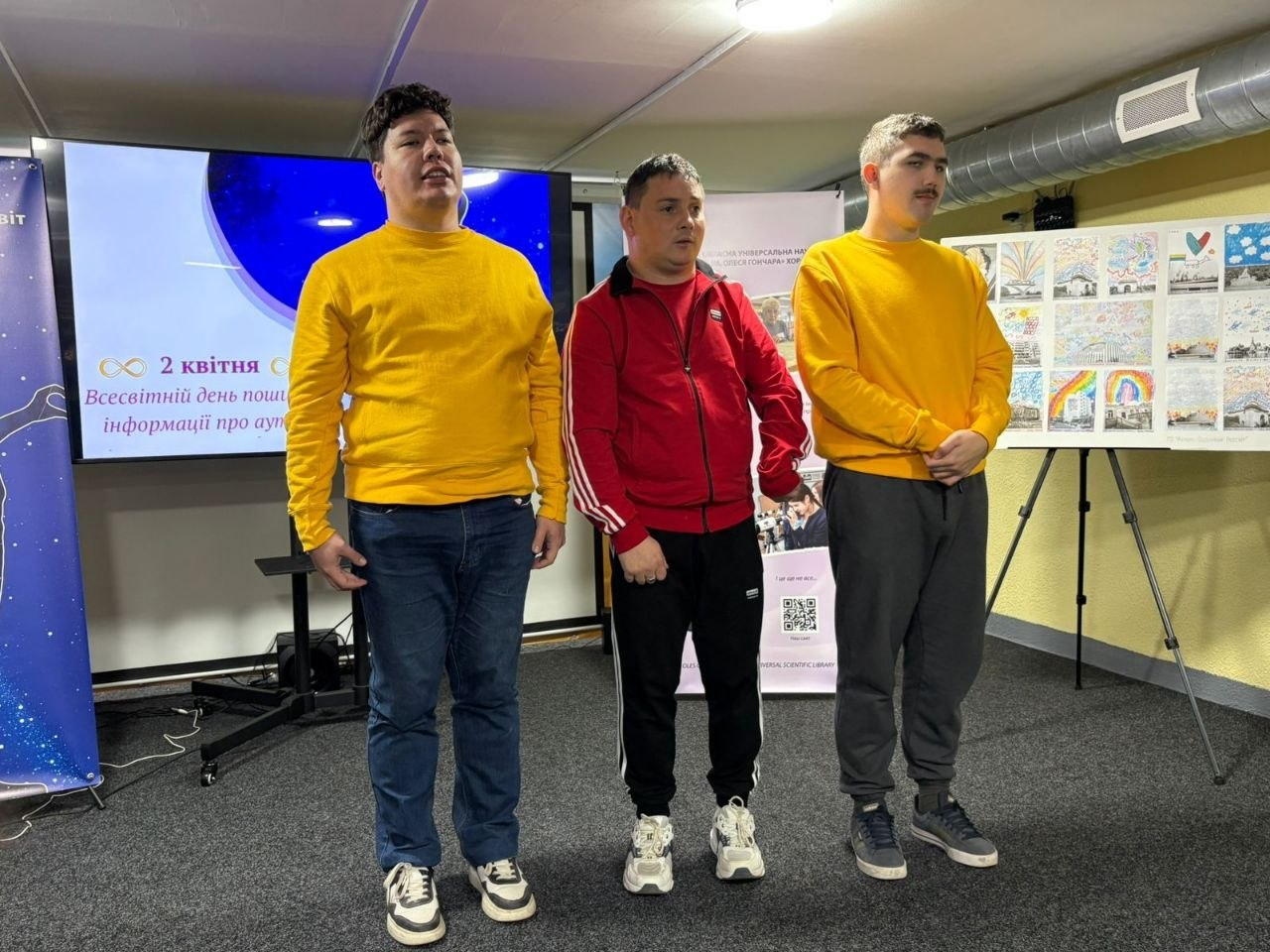 Троє молодих чоловіків стоять у приміщенні на фоні екрана з написом «2 квітня — Всесвітній день поширення інформації про аутизм». Праворуч від них розміщено мольберт із виставкою дитячих малюнків.