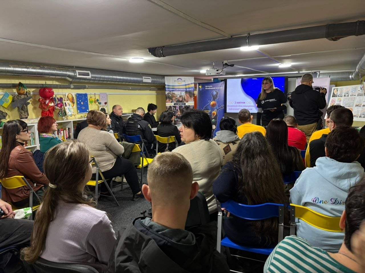 Слухачі в укритті під час заходу до Всесвітнього дня поширення інформації про аутизм. Люди сидять на стільцях перед екраном із презентацією. На фоні — книжкові полиці та дитячі малюнки.