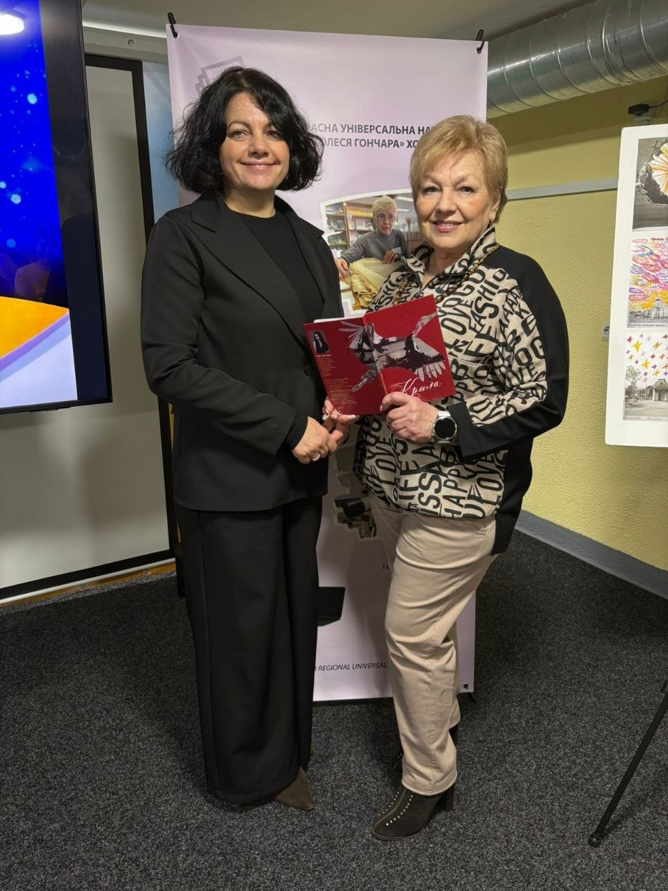 Дві усміхнені жінки у приміщенні бібліотеки тримають книгу з червоною обкладинкою. Вони стоять на фоні банера книгозбірні та мольберта з малюнками. Одна жінка одягнена в чорний костюм, інша — у принтовану блузу та світлі штани.