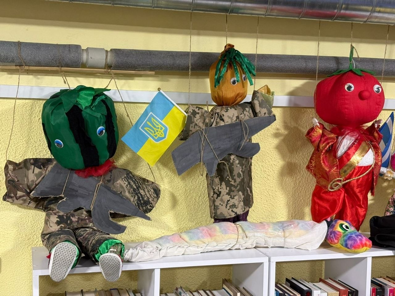 Три саморобні м'які ляльки у вигляді овочів — кавуна, гарбуза та помідора — стоять на книжковій полиці. Ляльки-овочі одягнені у військову форму або національне вбрання, тримають макети зброї та прапор України.