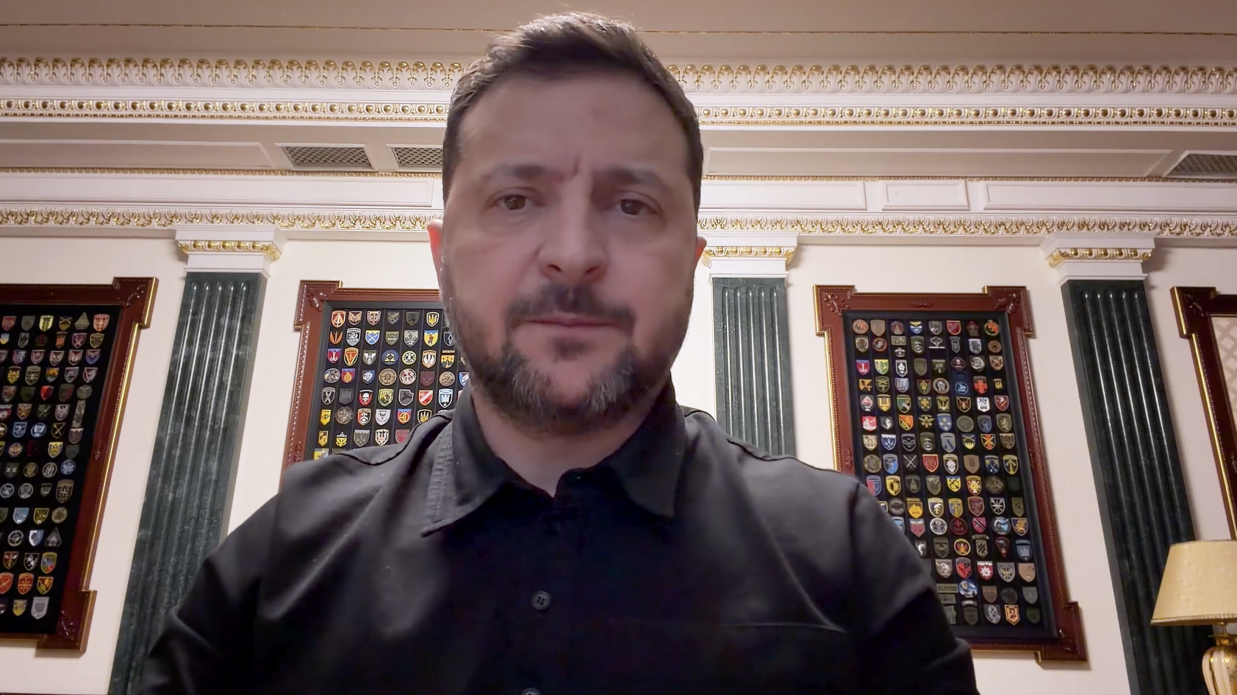 Чоловік у чорній сорочці на фоні кабінету з класичним інтер'єром. Позаду нього на стінах розміщені стенди з численними військовими шевронами та емблемами підрозділів.