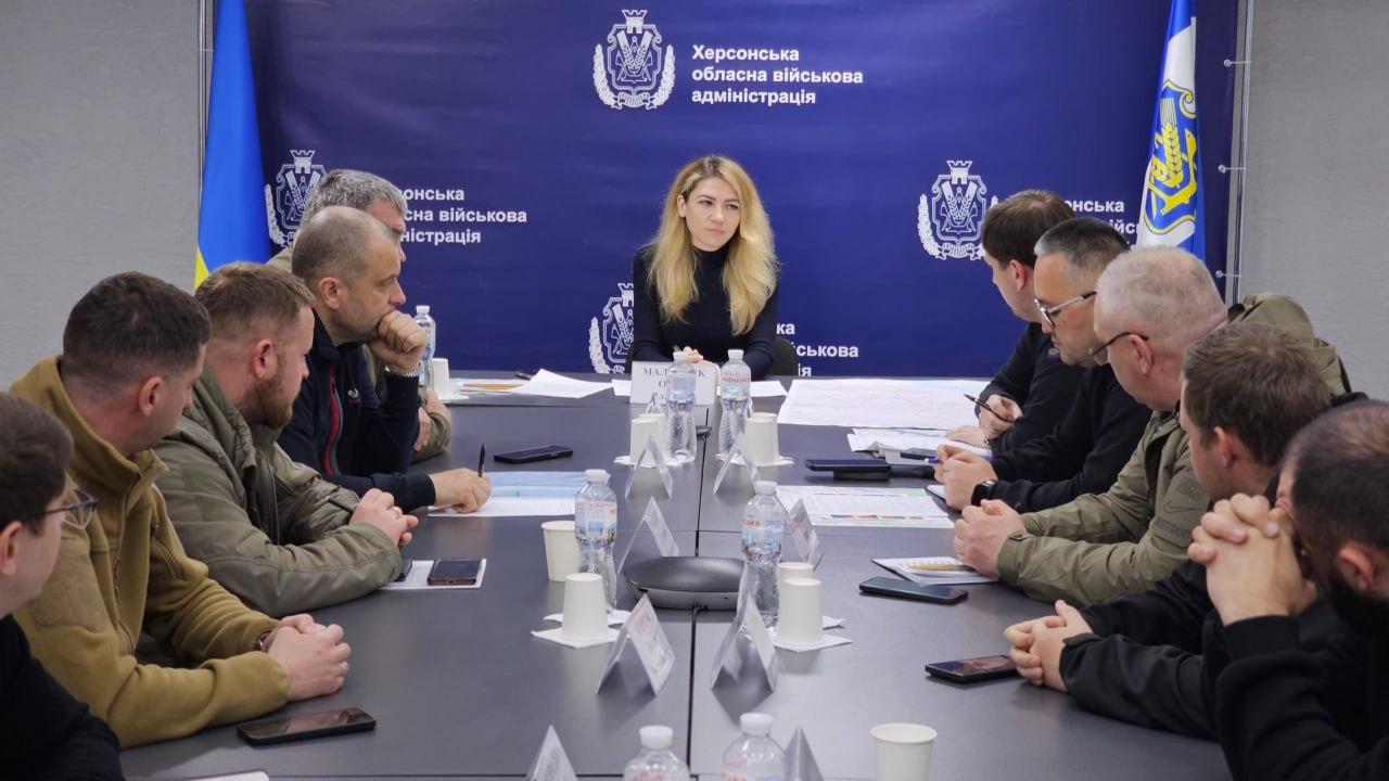Група людей у цивільному та військовому одязі сидить за великим столом під час наради в офіційному приміщенні на тлі банера Херсонської ОДА.