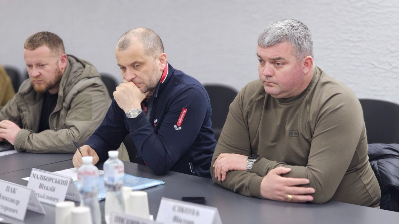 Троє чоловіків у повсякденному та тактичному одязі зосереджено сидять за столом під час робочої наради. На столі перед ними стоять пляшки з водою, склянки та таблички з іменами.