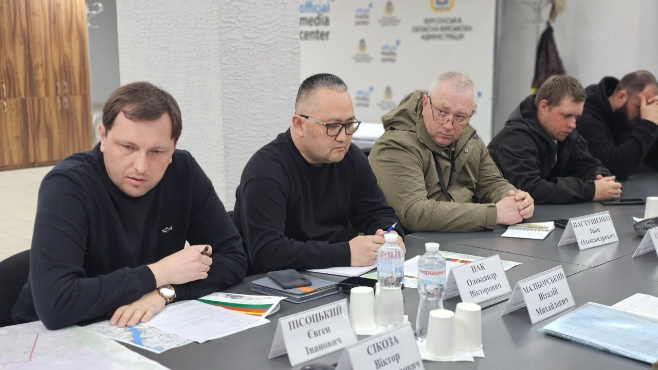 Група чоловіків у цивільному та тактичному одязі сидить за робочим столом під час офіційної зустрічі. Перед ними розкладені документи, карти, стоять пляшки з водою та настільні таблички. На задньому плані видно світлу стіну з логотипами офіційного медіацентру.