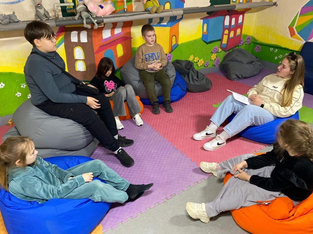 Група дітей та молода жінка спілкуються, сидячи на різнокольорових кріслах-мішках у яскравій ігровій кімнаті. Стіни прикрашені барвистими малюнками, що створює затишну атмосферу для заняття чи відпочинку.