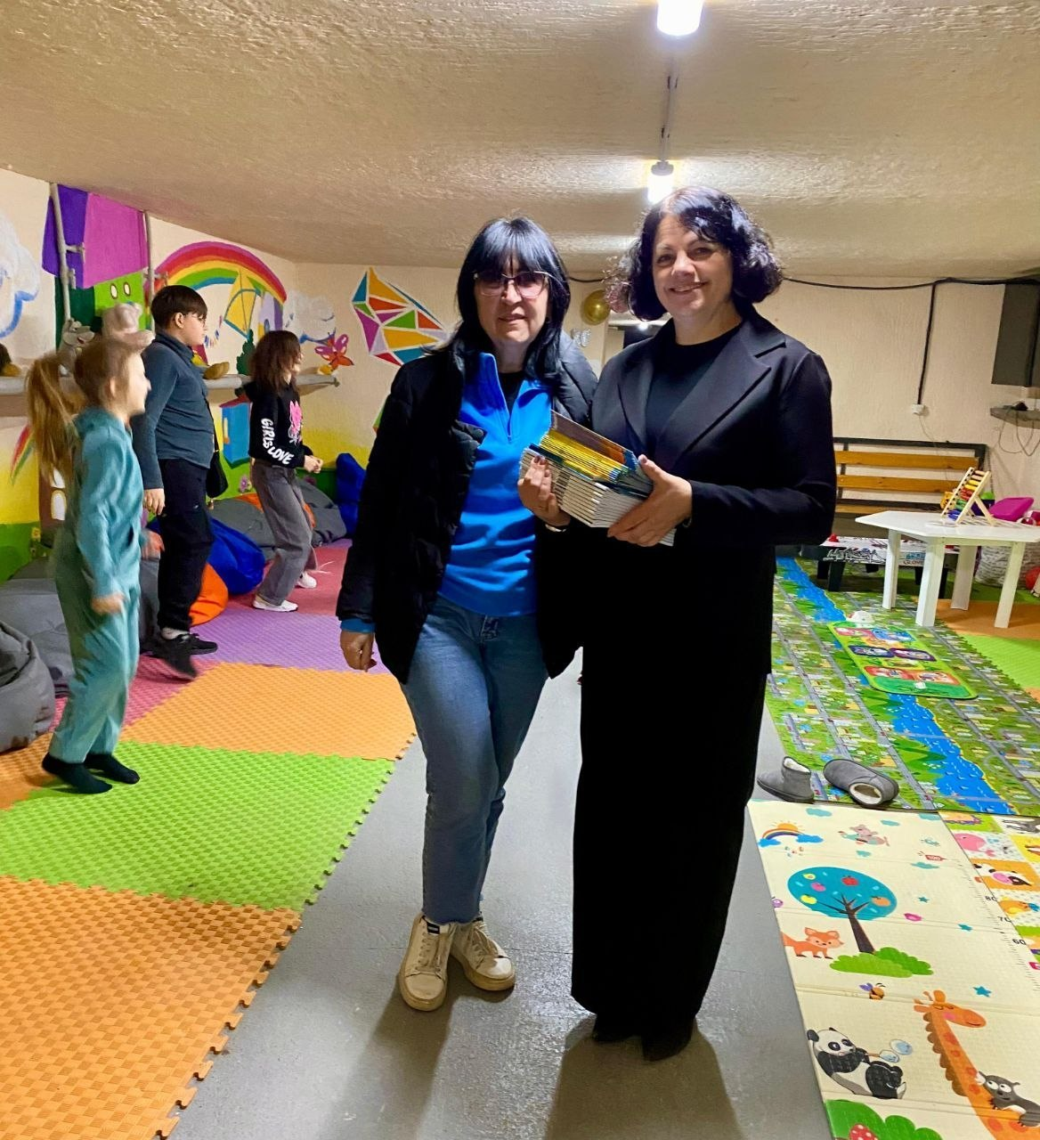 Дві жінки стоять у центрі яскраво оформленої дитячої ігрової кімнати. Одна з них тримає стопку книг. На фоні видно дітей та кольорові м’які мати на підлозі, а стіни прикрашені барвистими малюнками.