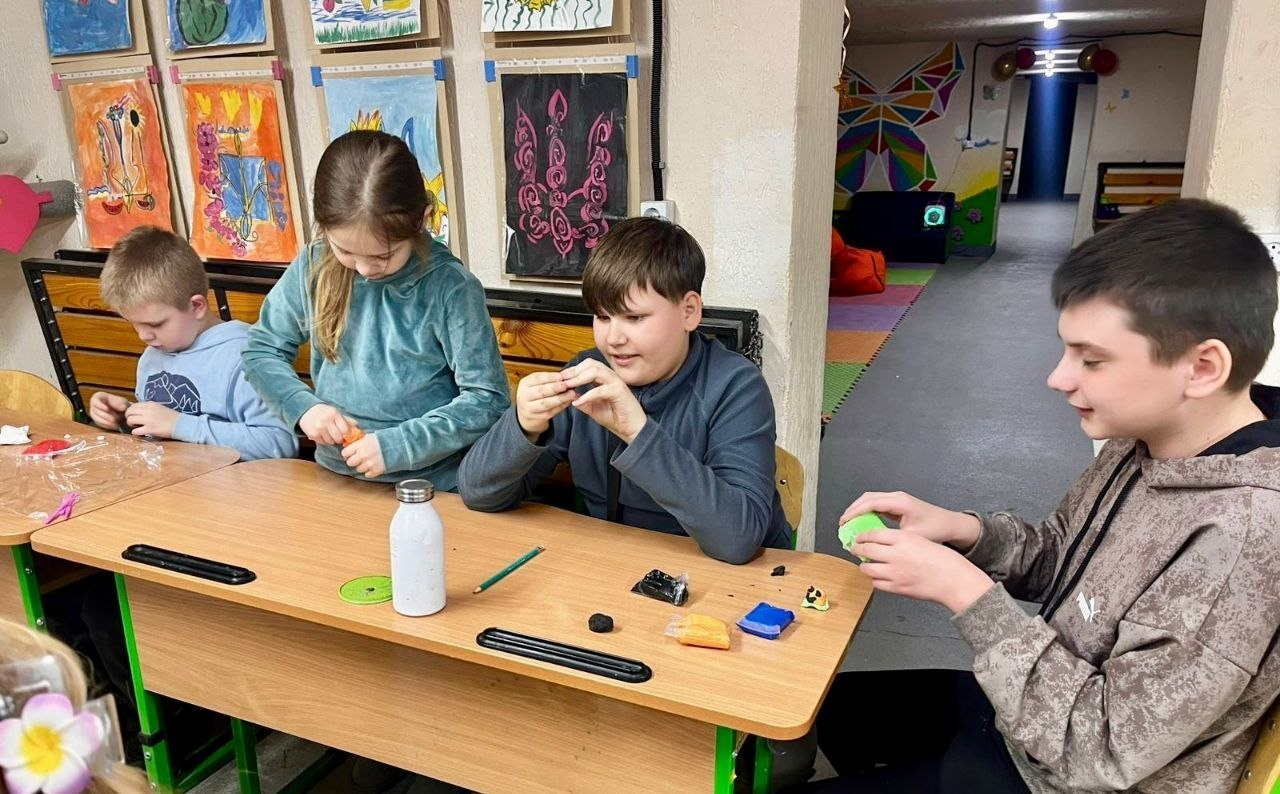 Четверо дітей сидять за довгим дерев’яним столом у творчій студії та захоплено ліплять фігурки з пластиліну. На стіні за ними розвішані дитячі малюнки, а на фоні видно ігрову зону з кольоровими декораціями.