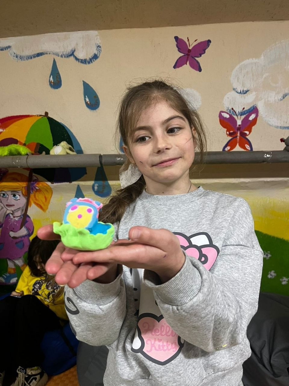 Дівчинка тримає на долонях маленьку яскраву фігурку, зліплену з пластиліну. Вона стоїть на фоні стіни з дитячими малюнками хмаринок, метеликів та парасольки.