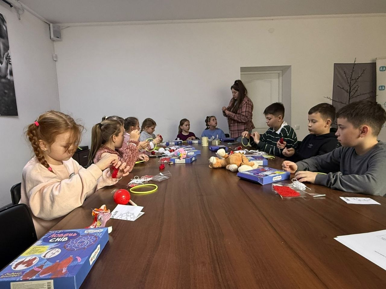 Група дітей сидить за довгим столом і виготовляє поробки під наглядом дорослої людини. На столі розкладені набори для творчості, нитки та іграшки, а діти зосереджено працюють руками.