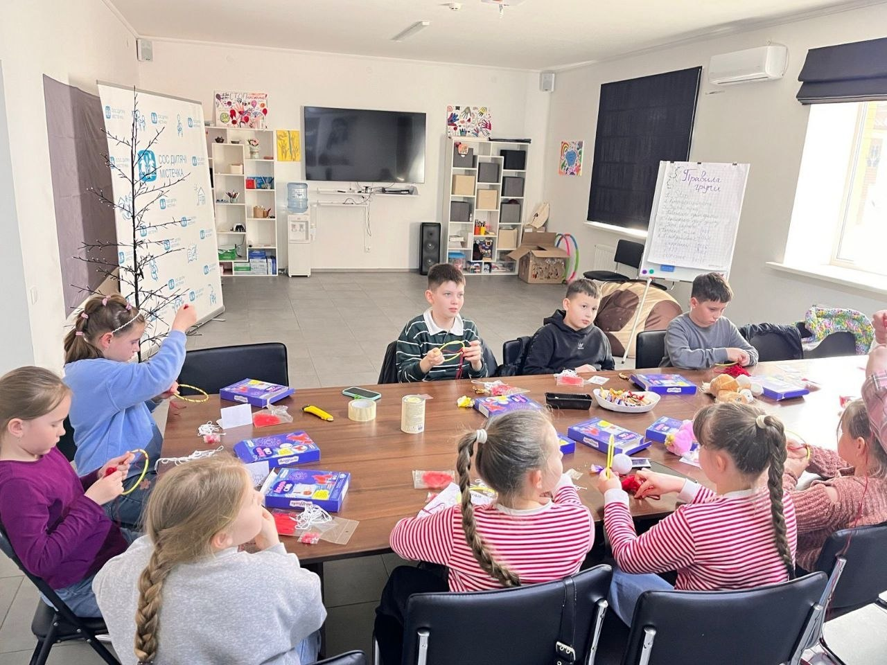 Група дітей сидить за великим столом у світлій кімнаті та займається рукоділлям. Вони зосереджено виготовляють поробки з ниток та кольорових матеріалів під час творчого заняття.