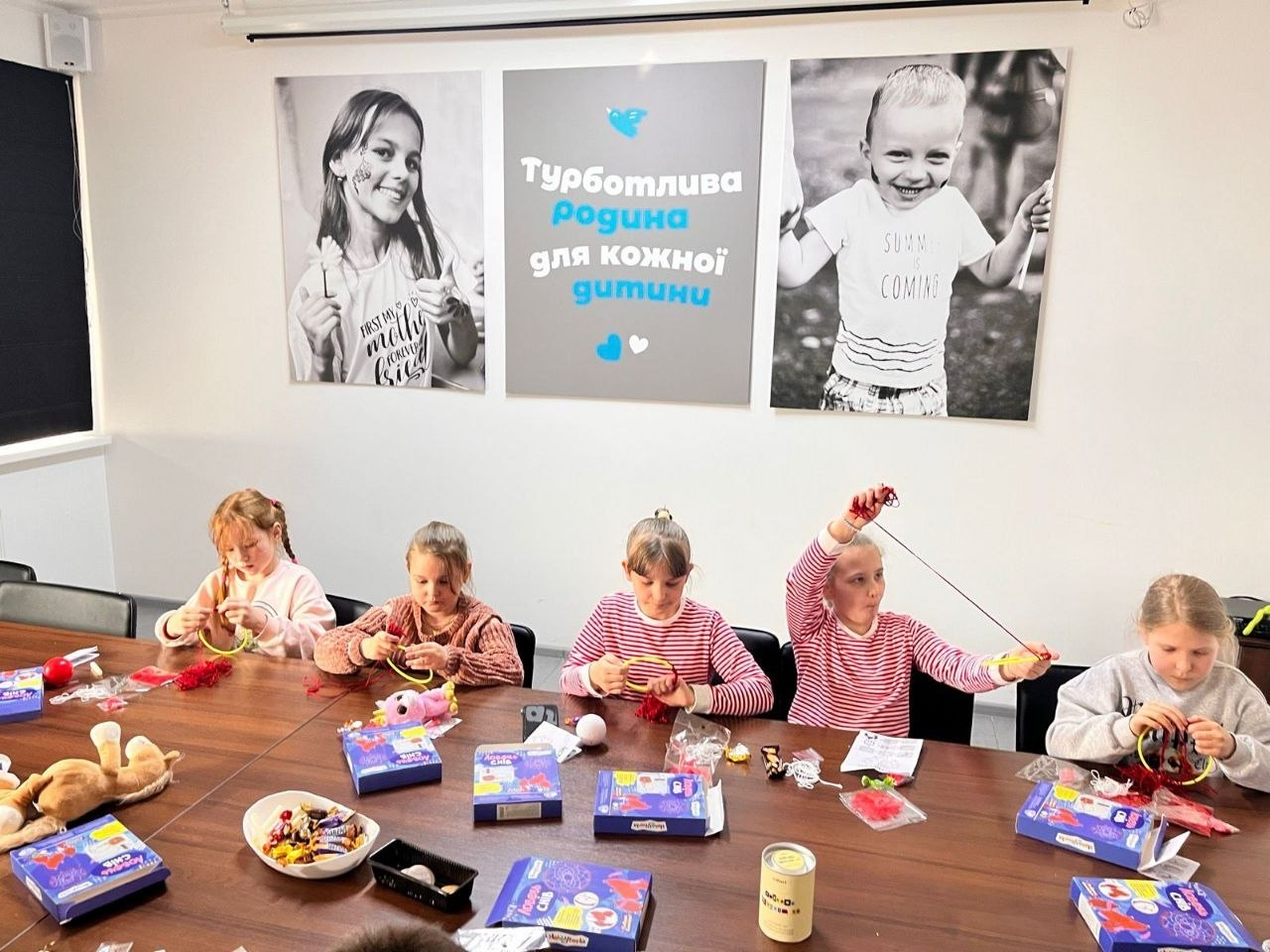 П'ятеро дівчаток сидять за довгим столом і виготовляють поробки з ниток та обручів. На стіні над ними розміщено великий напис «Турботлива родина для кожної дитини» та два чорно-білі фото усміхнених дітей.