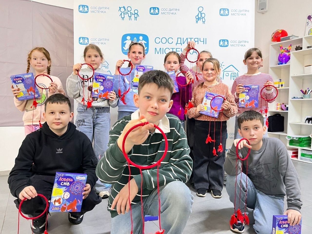 Група дітей стоїть і сидить на фоні банера, демонструючи виготовлені червоні ловці снів та набори для творчості. Вони посміхаються, тримаючи свої поробки перед собою в сучасно обладнаній кімнаті.