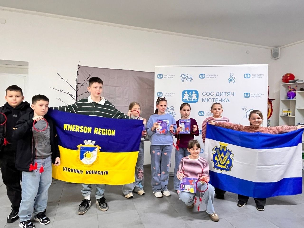 Група дітей у світлій кімнаті тримає прапори Херсонської області та селища Верхній Рогачик. Поруч розташований банер з логотипом «СОС Дитячі Містечка», а діти демонструють свої творчі поробки.