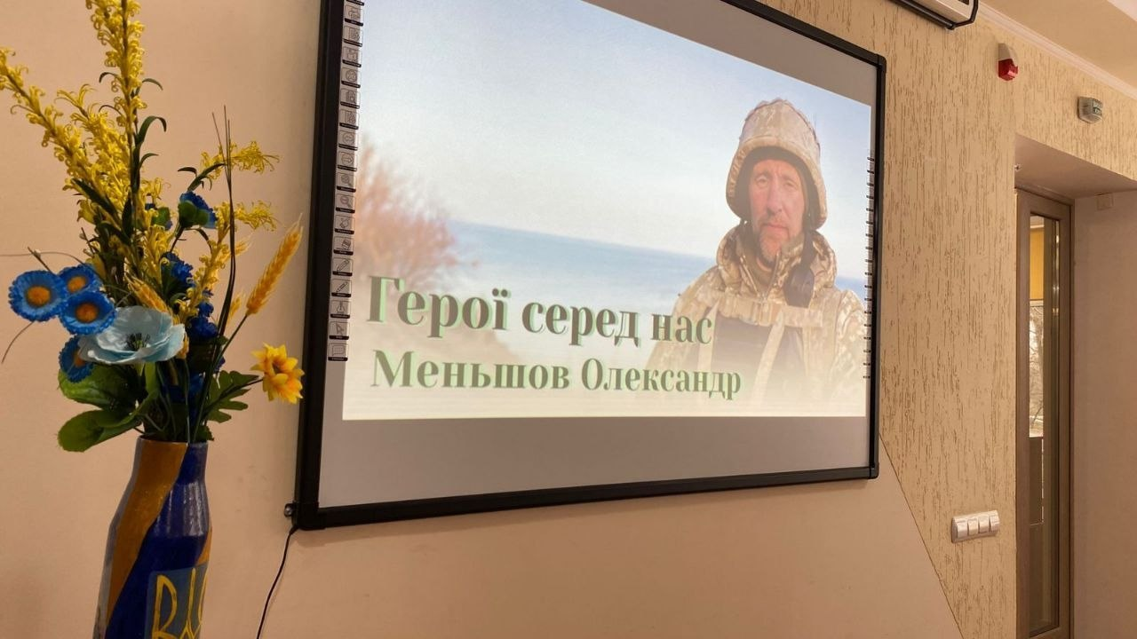 Проєкційний екран на стіні у світлому приміщенні. На екрані відображено портрет військового Олександра Меньшова з написом «Герої серед нас». Ліворуч від екрана стоїть патріотична композиція з квітів у синьо-жовтих кольорах.