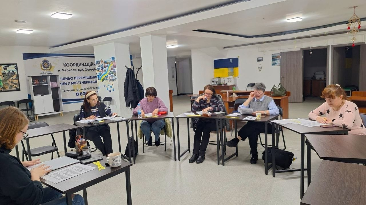Група жінок сидить за столами, розставленими півколом у світлому приміщенні. На стіні позаду видно банер з українською символікою та написом «Координаційний центр».