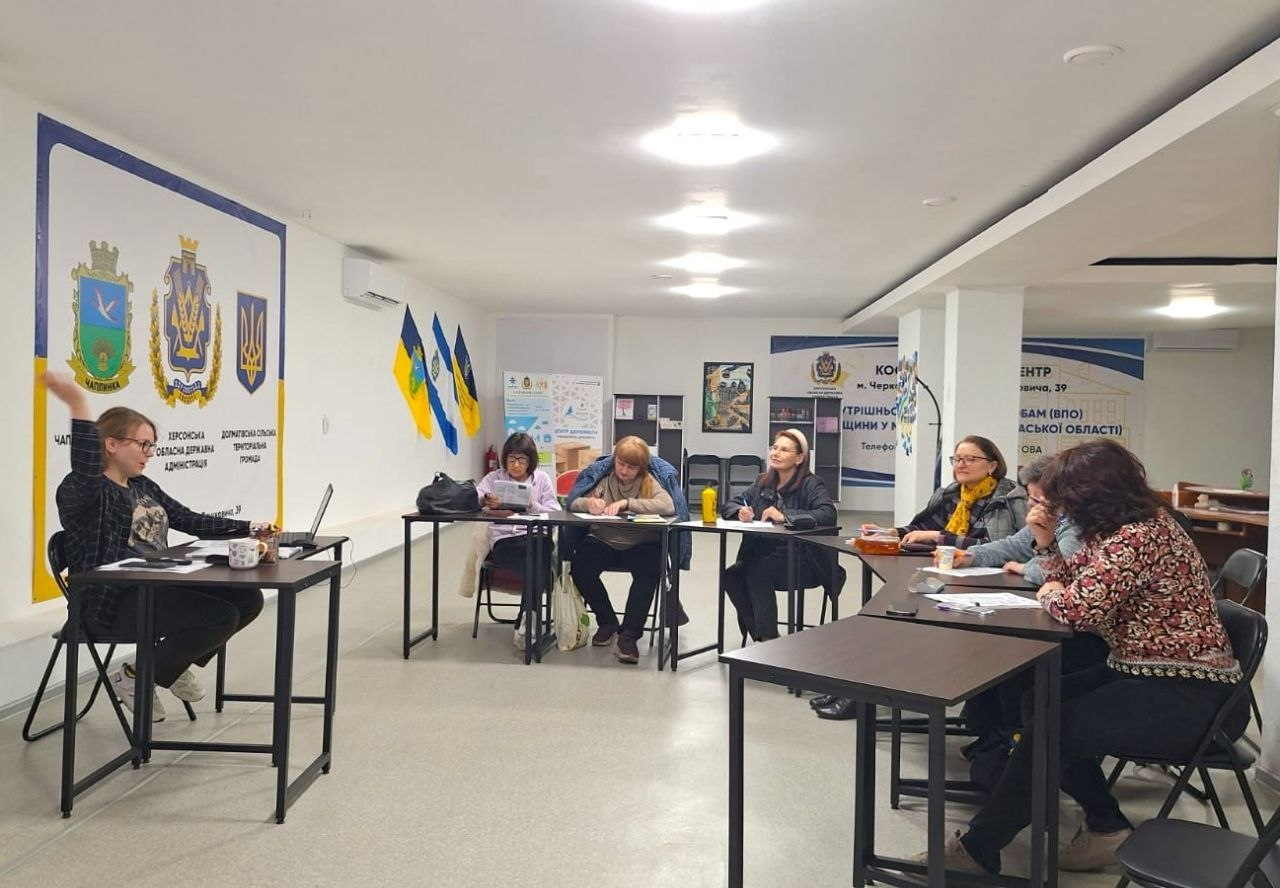 Група людей сидить за столами, розставленими півколом у світлій залі, та бере участь у робочій зустрічі. На стінах розміщено інформаційні плакати з українською символікою та державні прапори.