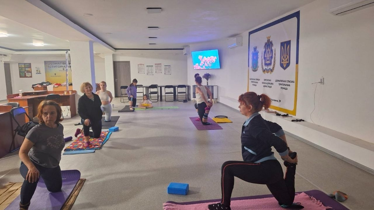 Група жінок займається фітнесом або розтяжкою у просторому світлому приміщенні. Вони виконують вправи на килимках, використовуючи спеціальні блоки. На стіні залу розміщено банер із державною символікою України та інформаційні плакати.