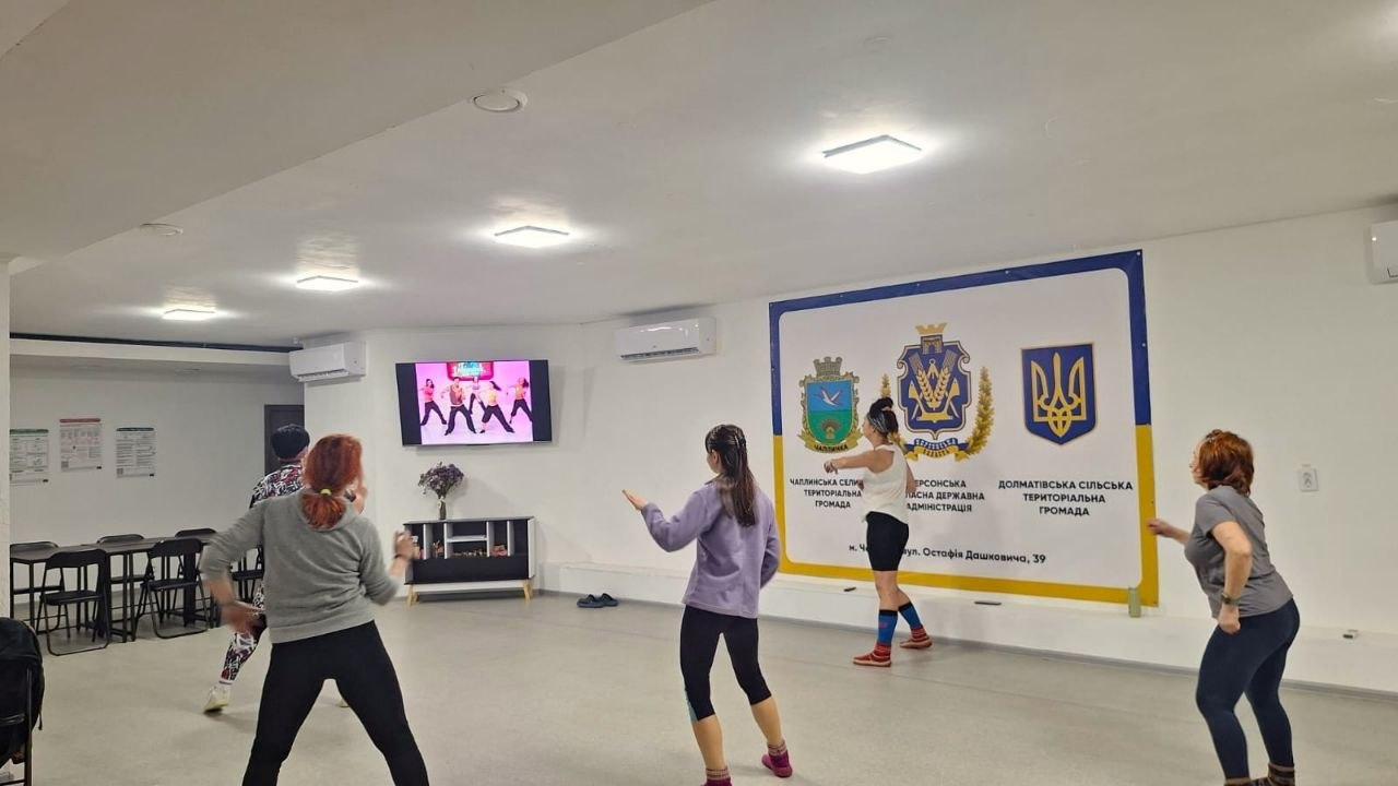 Група жінок займається танцями або фітнесом у просторому світлому залі, повторюючи рухи за відео на моніторі. На стіні розміщено великий інформаційний банер із державною символікою та гербами територіальних громад.
