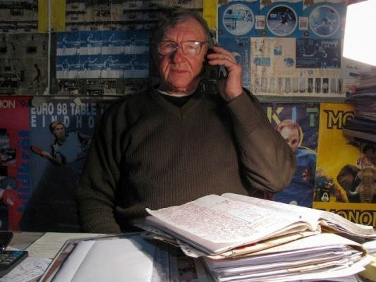 Чоловік в окулярах та темному светрі розмовляє по мобільному телефону, сидячи за столом із великою кількістю паперів та папок. Стіни за ним обклеєні плакатами та вирізками на спортивну тематику.