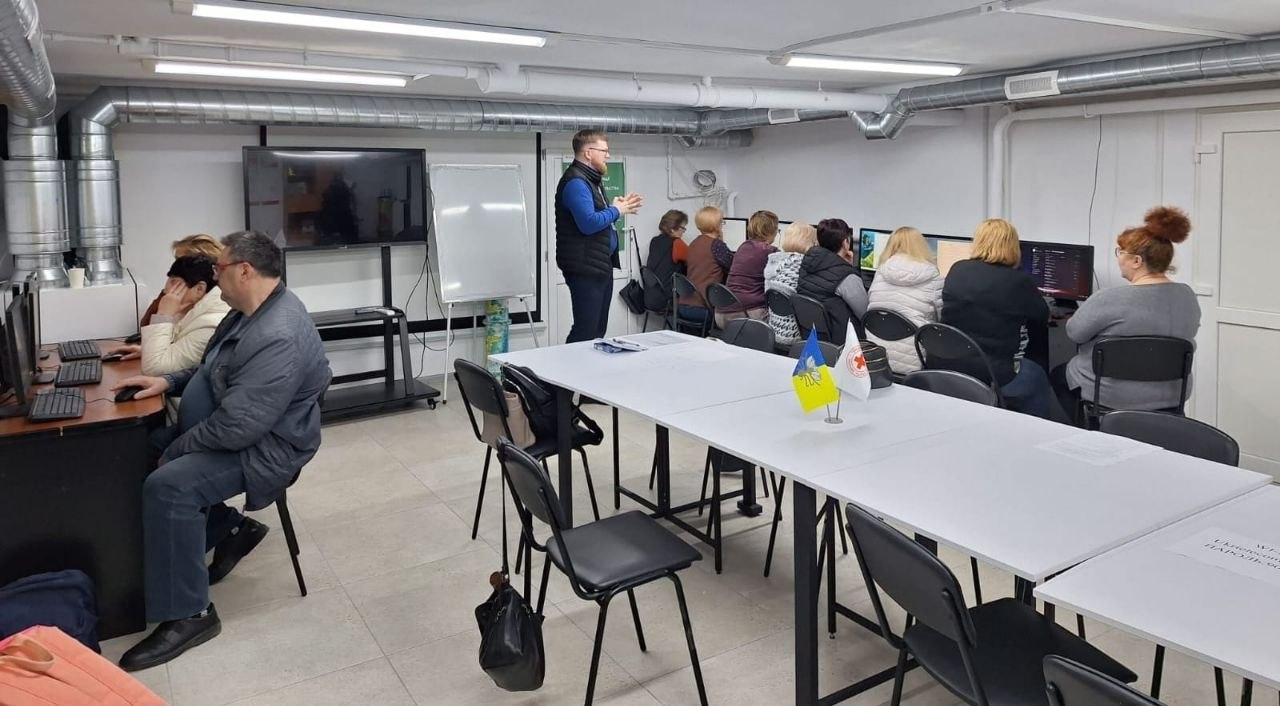 Група людей займається за комп'ютерами в облаштованому навчальному класі або коворкінгу. Викладач стоїть біля дошки, а на передньому плані — довгий стіл із прапорцями України та Червоного Хреста.