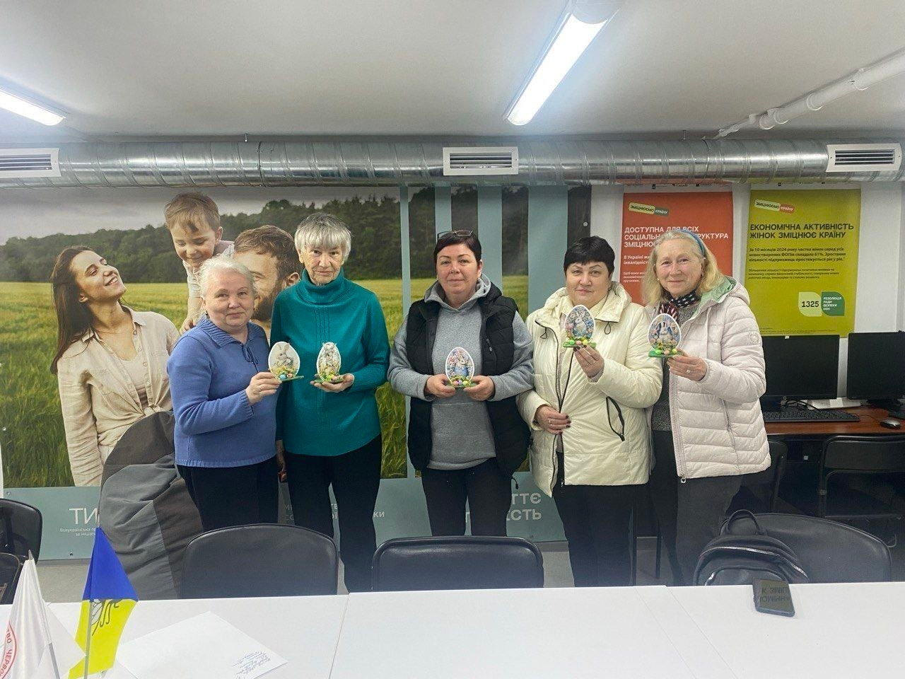 Група жінок у приміщенні тримає в руках декоративні великодні яйця. На задньому плані — інформаційні плакати, а на столі попереду стоять маленькі прапорці України та Червоного Хреста.