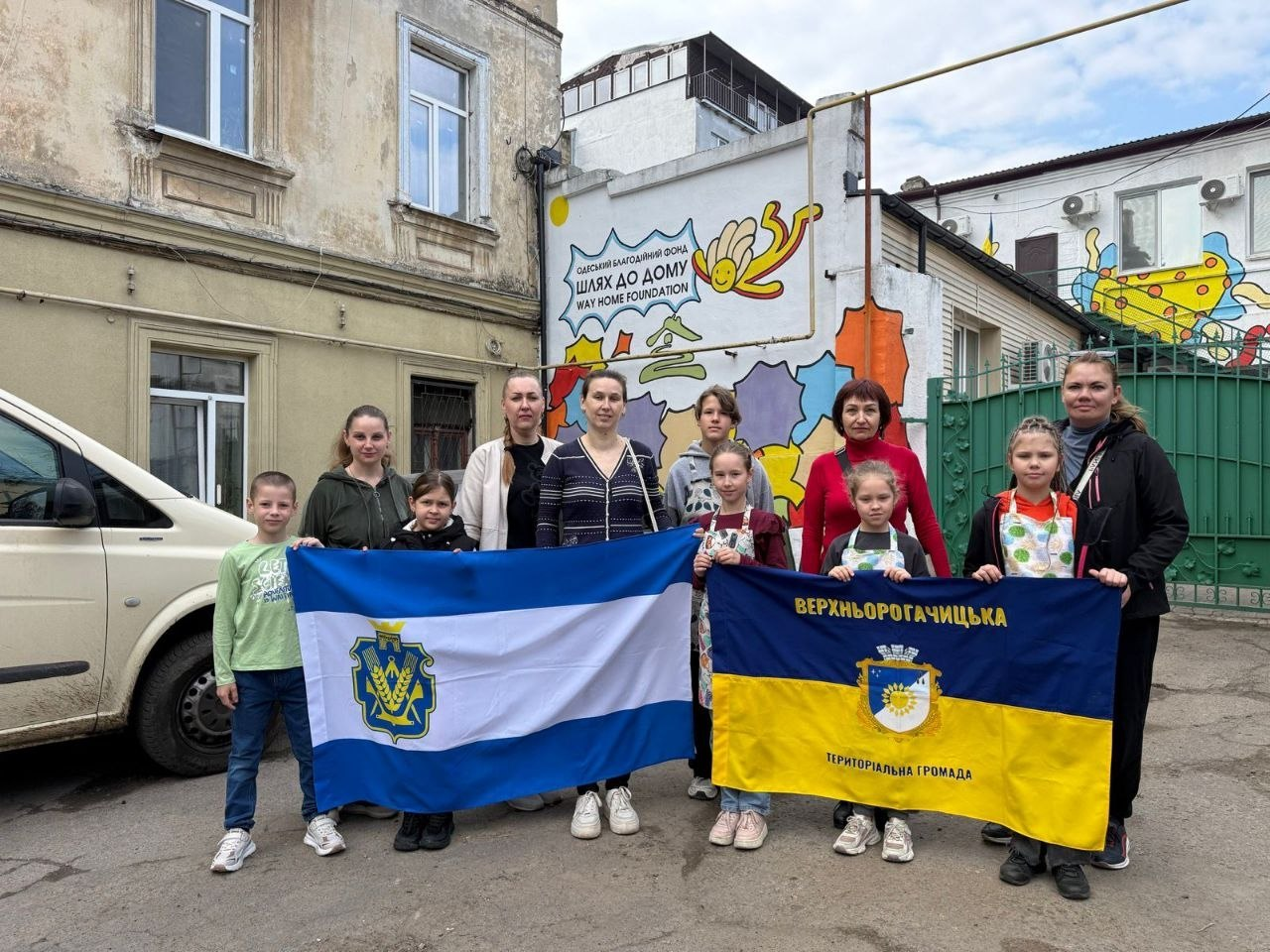 Група дорослих та дітей позує на вулиці, тримаючи прапори міста Херсон та Верхньорогачицької територіальної громади. Вони стоять на фоні будівлі з муралом благодійного фонду «Шлях до дому».