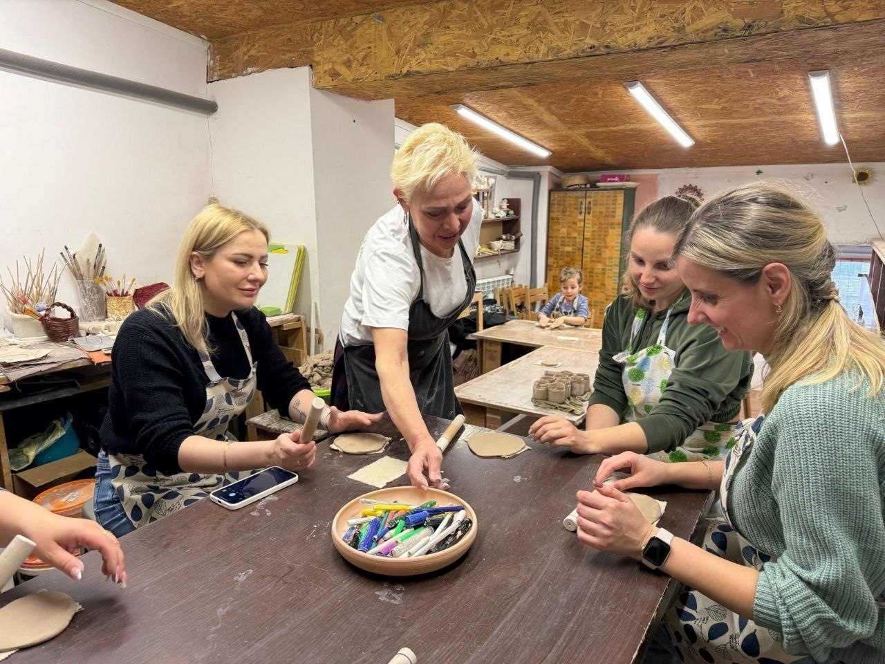 Жінки в фартухах працюють з глиною за великим столом у гончарній майстерні. Одна з учасниць допомагає іншим, у центрі столу стоїть тарілка з фломастерами.