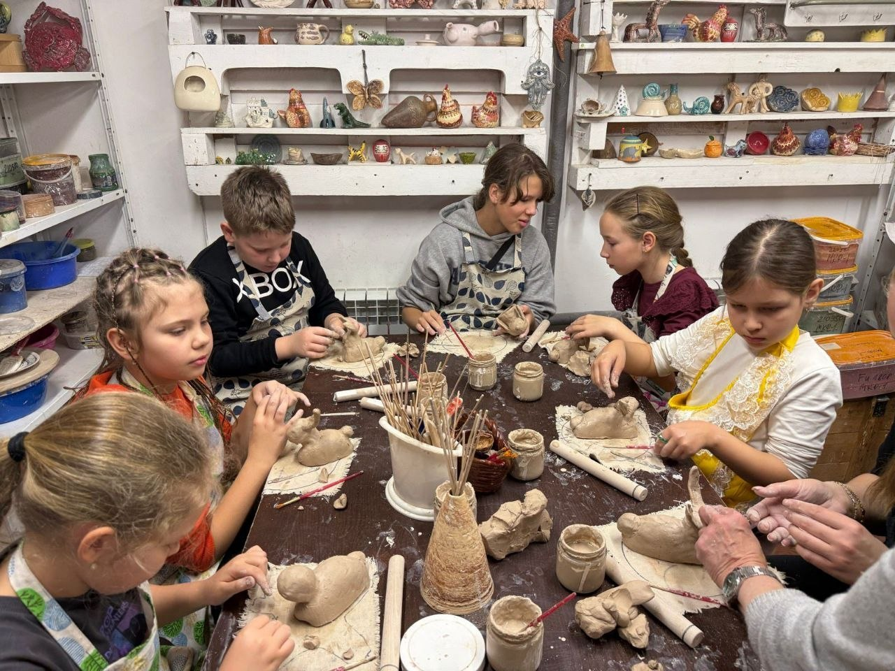 Група дітей у фартухах займається ліпленням із глини за великим столом у творчій майстерні. На полицях позаду виставлено багато готових керамічних виробів та фігурок.