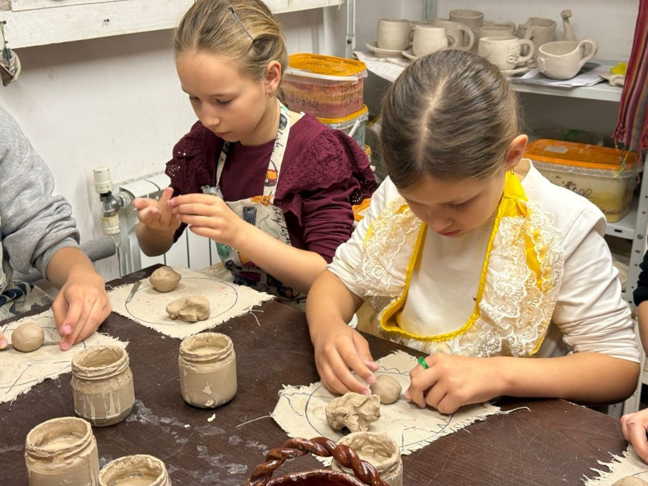 Дві дівчинки у фартухах зосереджено ліплять вироби з глини за столом у гончарній майстерні. На фоні видно полиці з готовими керамічними чашками та ємностями з матеріалами.