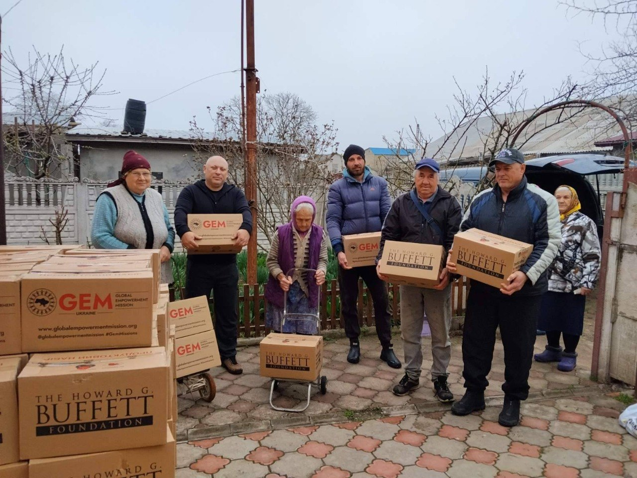 Група людей на подвір'ї тримає коробки з гуманітарною допомогою від фондів GEM та Howard G. Buffett Foundation. Поруч стоїть стос аналогічних картонних коробок.