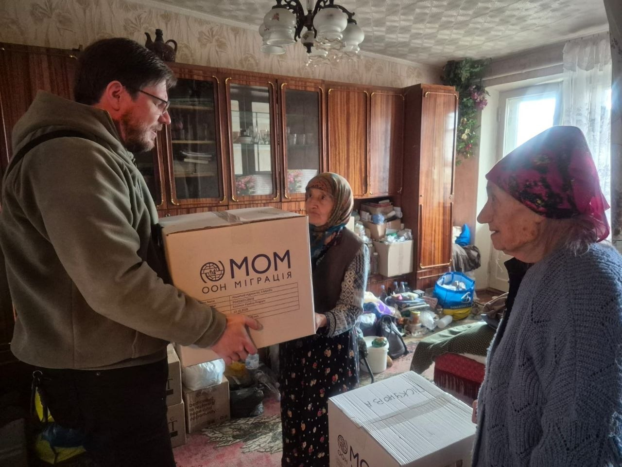 Волонтер передає коробку з логотипом «IOM ООН Міграція» літній жінці в її помешканні.