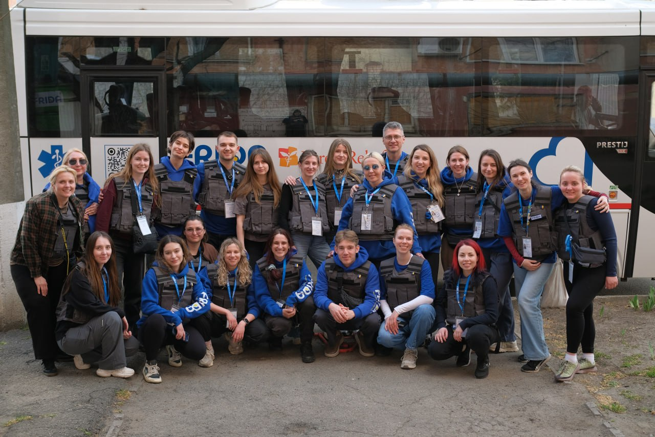 Група волонтерів у синьому одязі та захисних жилетах позує для спільного фото на фоні евакуаційного автобуса з логотипами медичних та гуманітарних організацій.