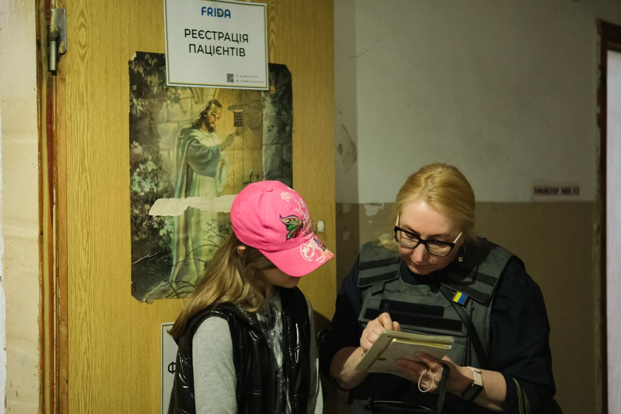 Медичний працівник у захисному жилеті робить записи в блокноті поруч із дівчинкою в рожевій кепці біля дверей із написом «Реєстрація пацієнтів».